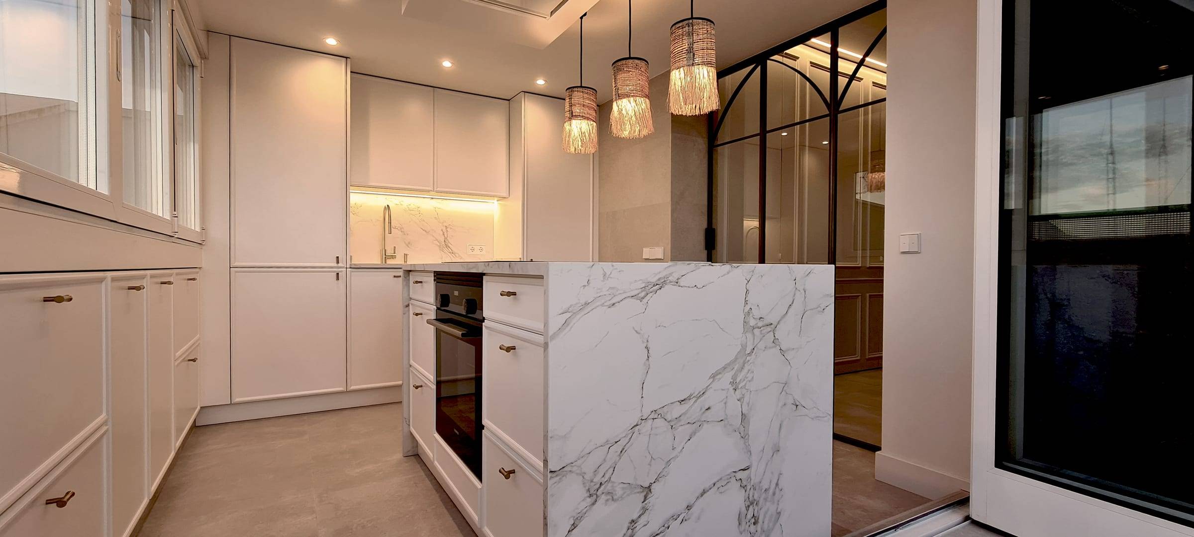 Cocina reformada con isla de acabado mármol y cerramiento de cristal hacia el salón