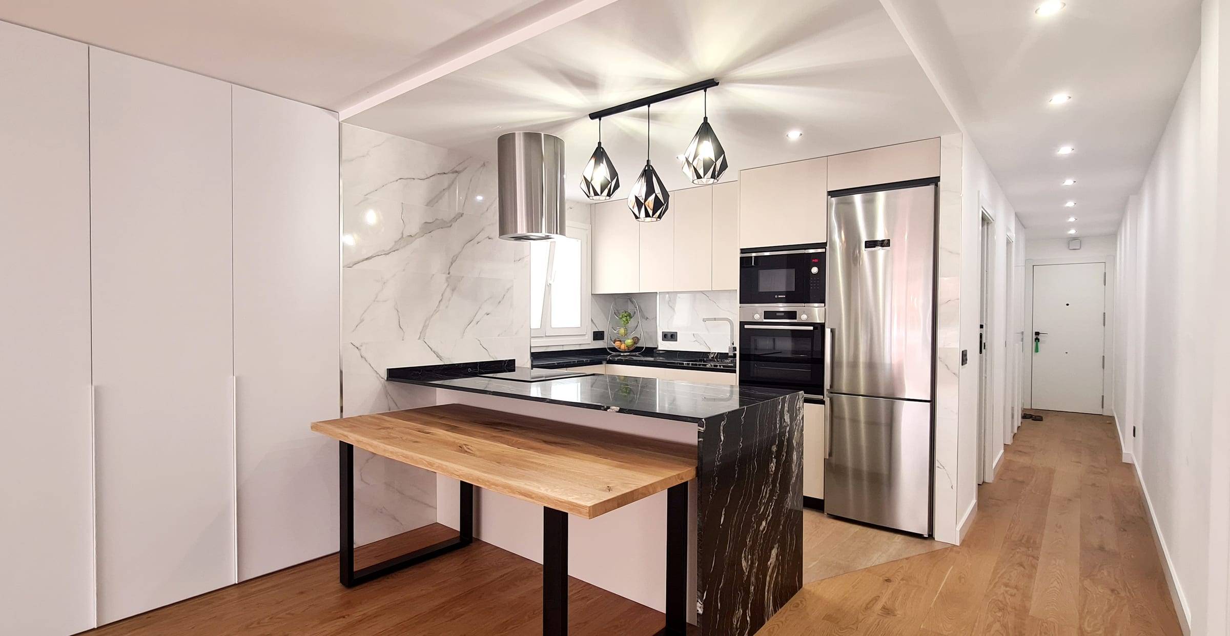 Cocina reformada con isla de encimera negra, mesa de madera y lámparas colgantes