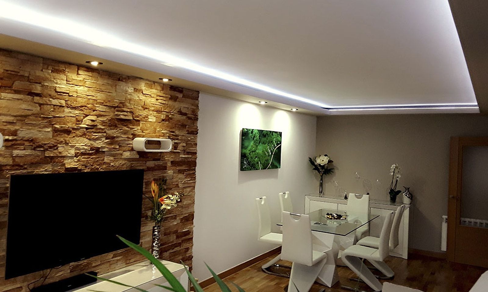 Salón con pared de piedra, tele colgada y moldura de techo con luz indirecta