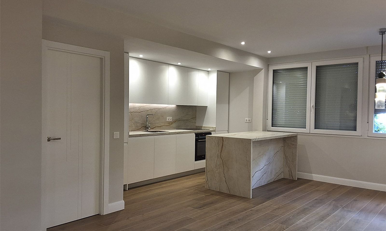 Salón comedor abierto a la cocina blanca con isla de mármol, suelo de imitación madera y ventana