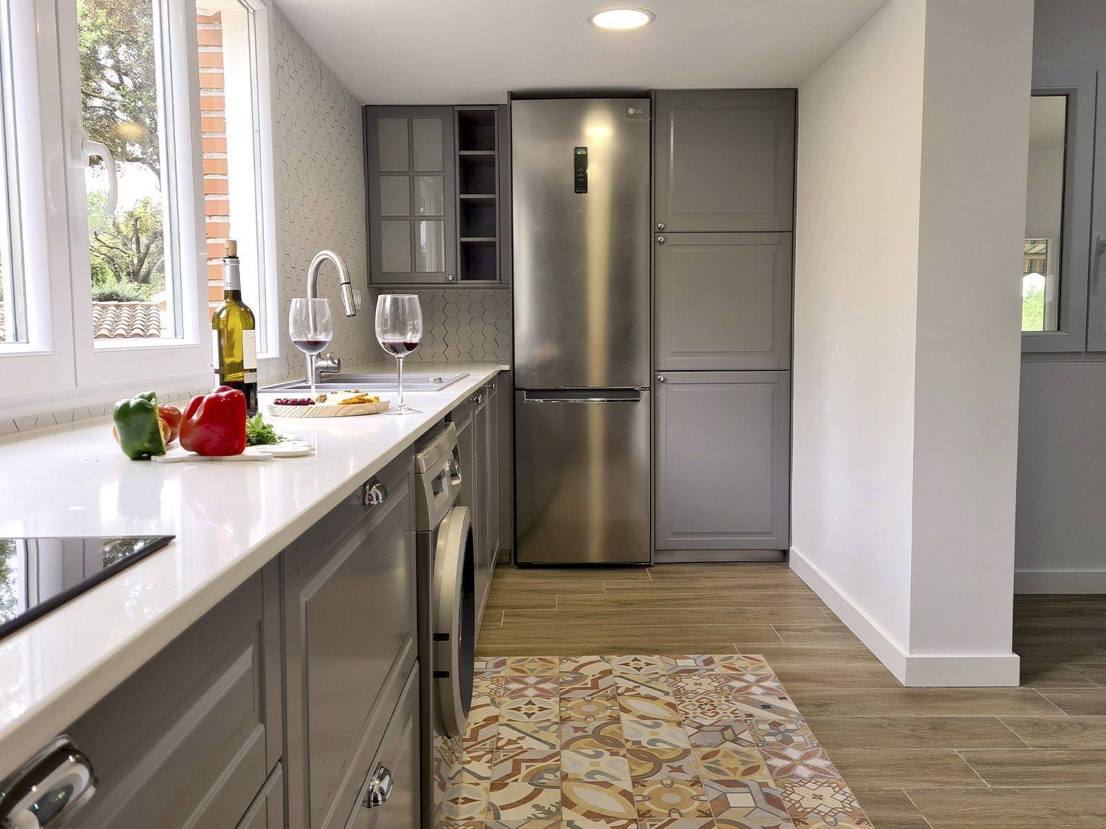 Cocina con muebles grises, frigorífico de acero, encimera blanca y ventana al jardín