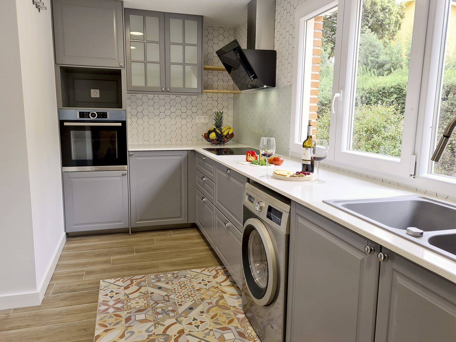 Cocina en L con muebles grises de cuarterones, horno en columna, lavadora y ventanas al jardín