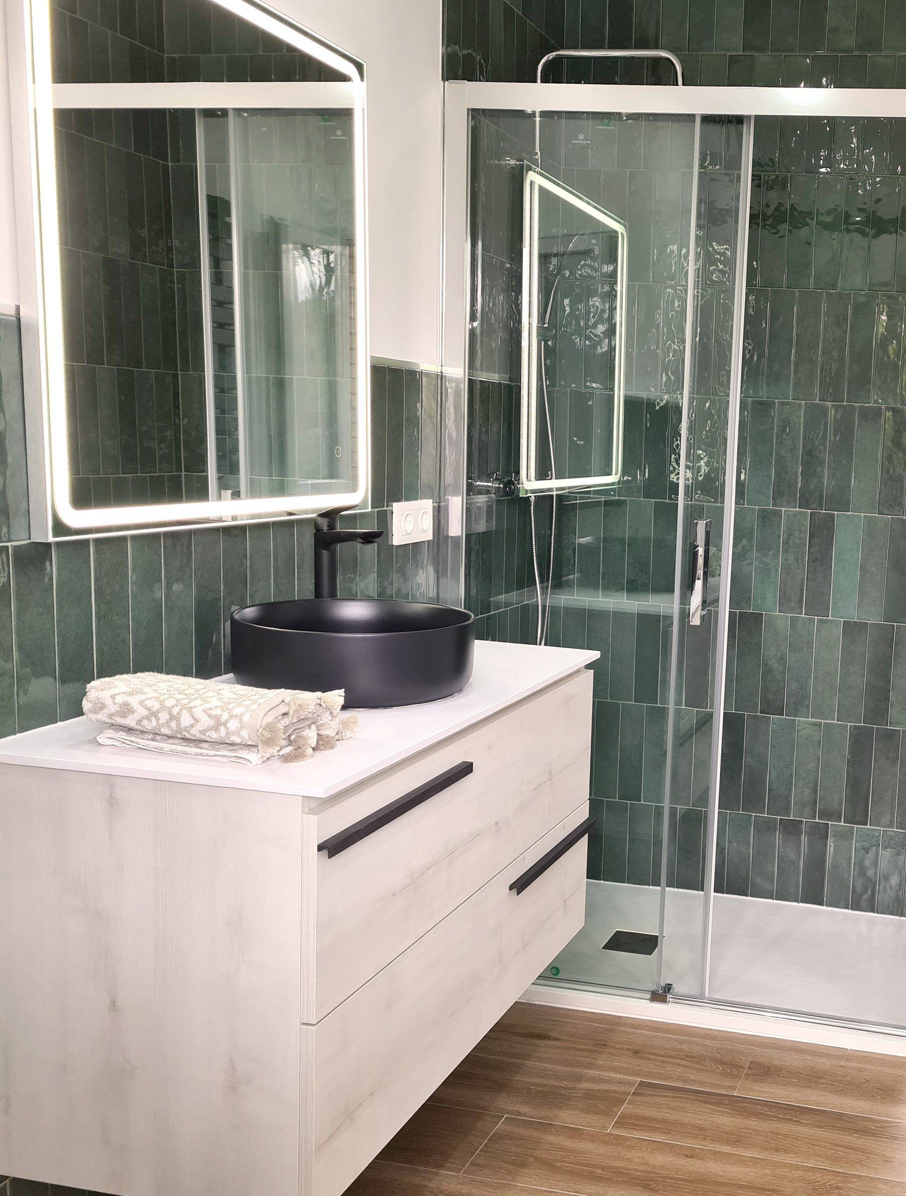 Baño con azulejo verde en vertical, mueble suspendido blanco, lavabo negro sobre encimera y mampara