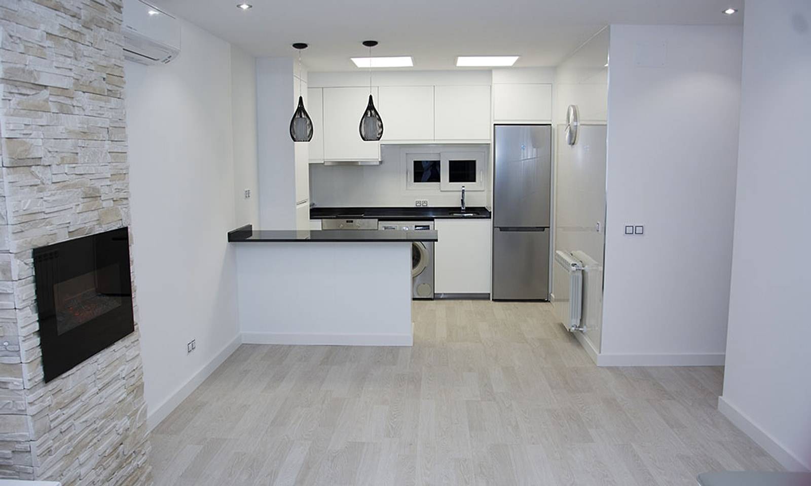 Cocina blanca abierta con barra de granito negro, lámparas colgantes y una chimenea en la pared de piedra