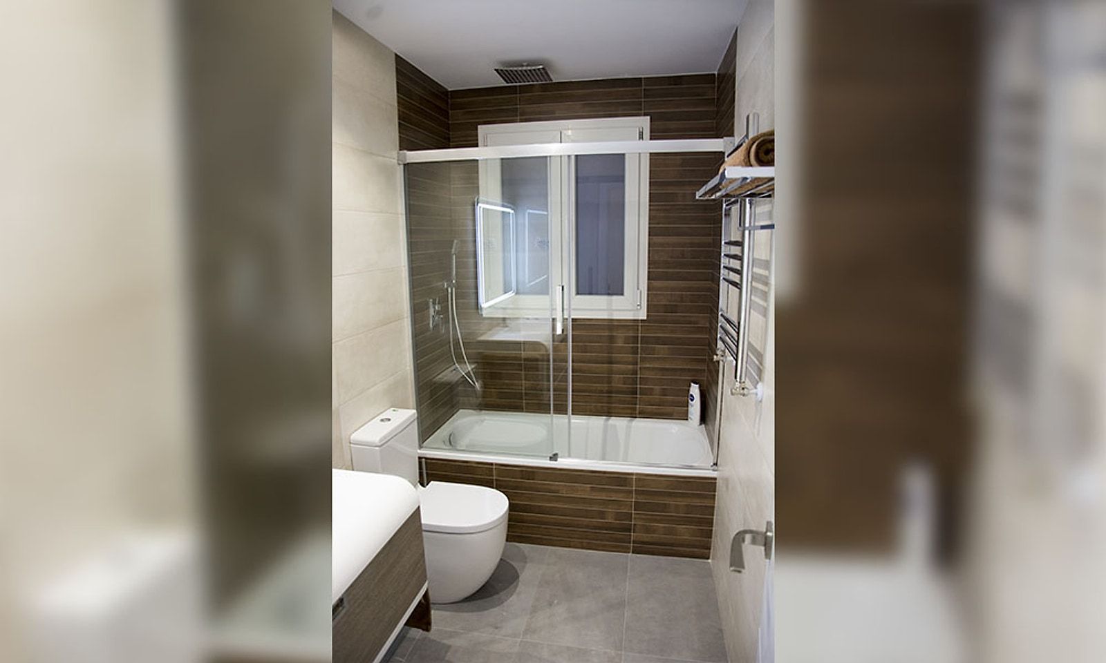 El mismo baño reformado, con bañera y mampara, alicatado claro y una franja de madera oscura