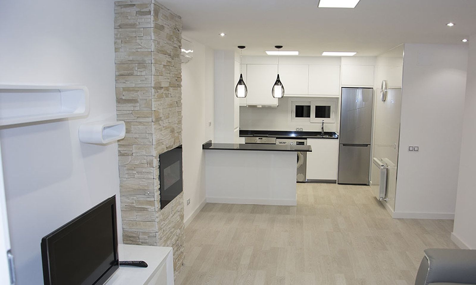 El mismo espacio terminado, con la cocina blanca abierta al salón y una pared de piedra