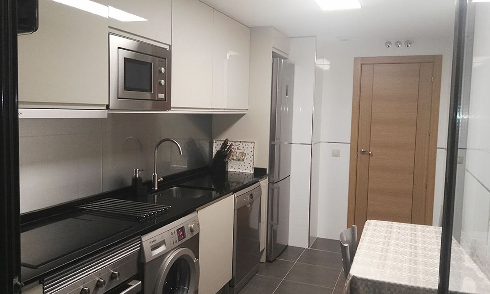 La misma cocina terminada, con muebles blancos, encimera negra y electrodomésticos de acero