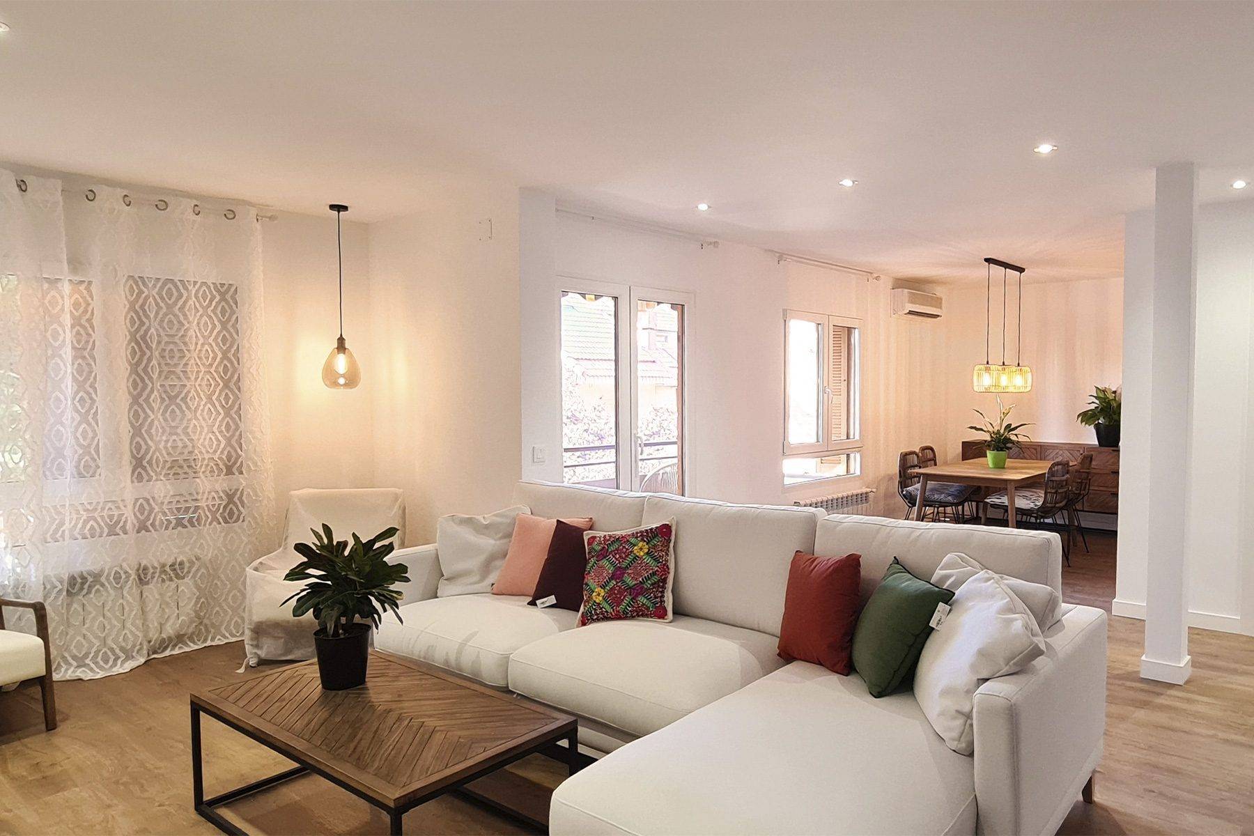 Salón amplio con sofá blanco, mesa baja de madera y el comedor al fondo, con suelo de madera y paredes blancas