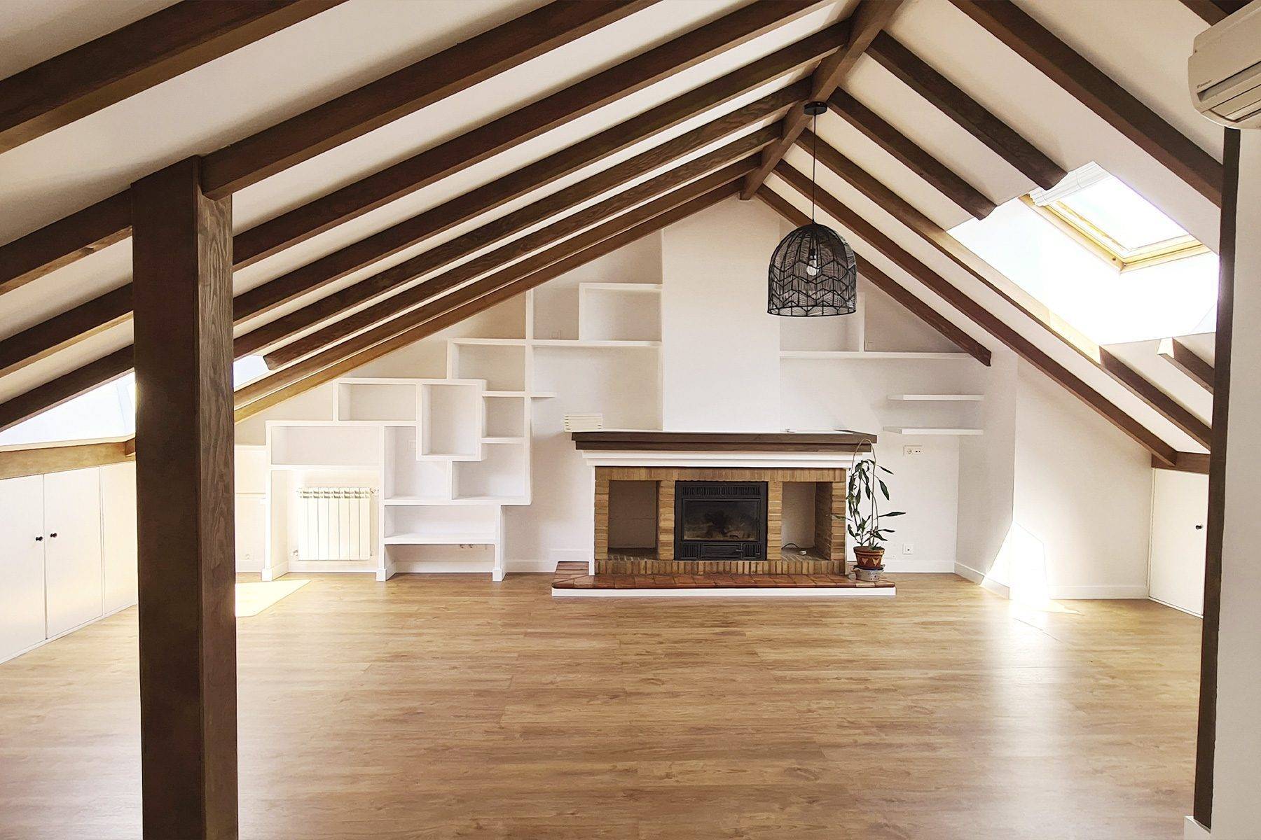 Planta bajo cubierta con vigas de madera vistas, chimenea, estantería blanca de obra y suelo de madera