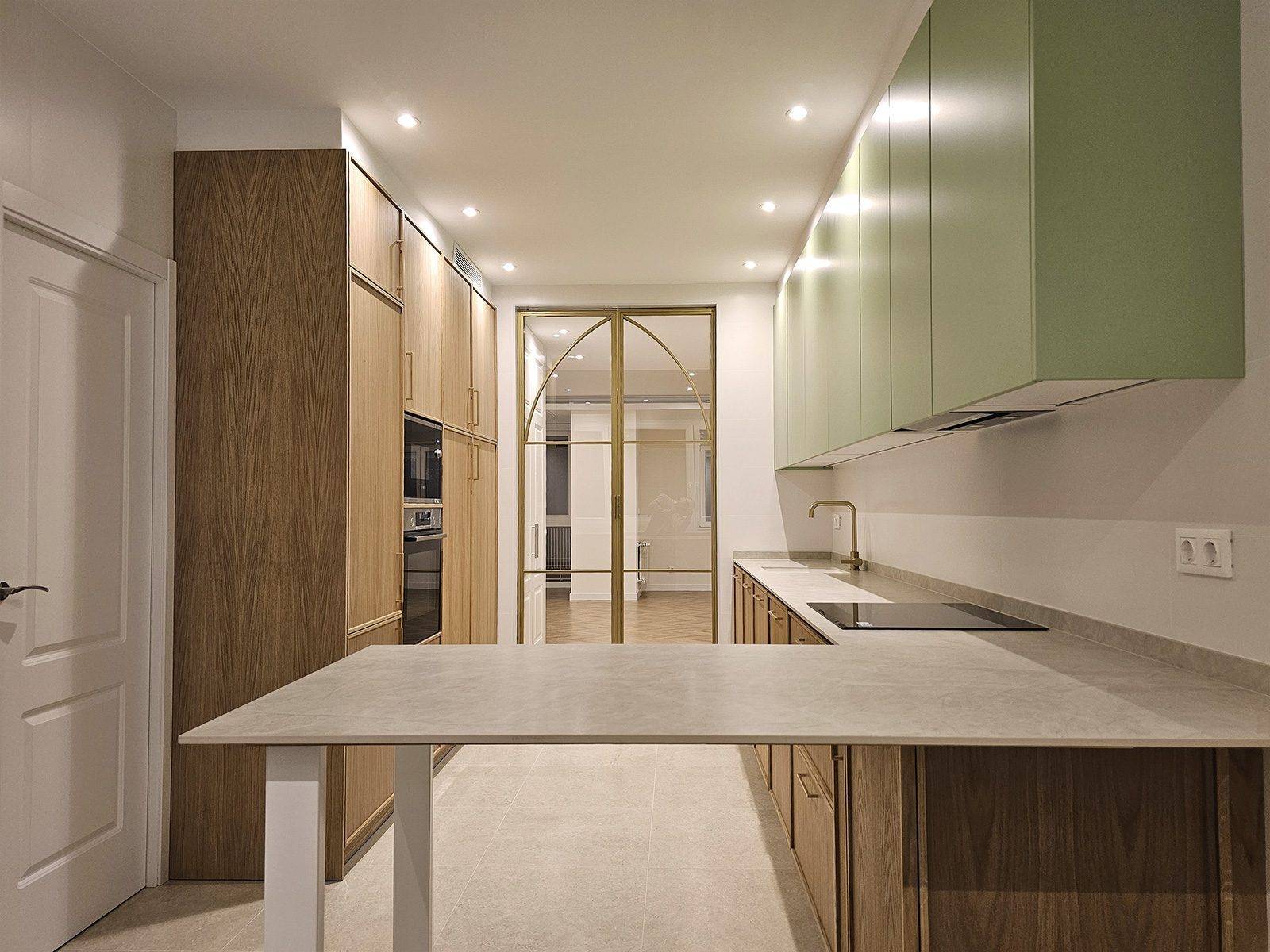 Cocina con columnas de madera, muebles altos verdes, península de encimera gris y puerta de cristal con perfil dorado en arco al fondo