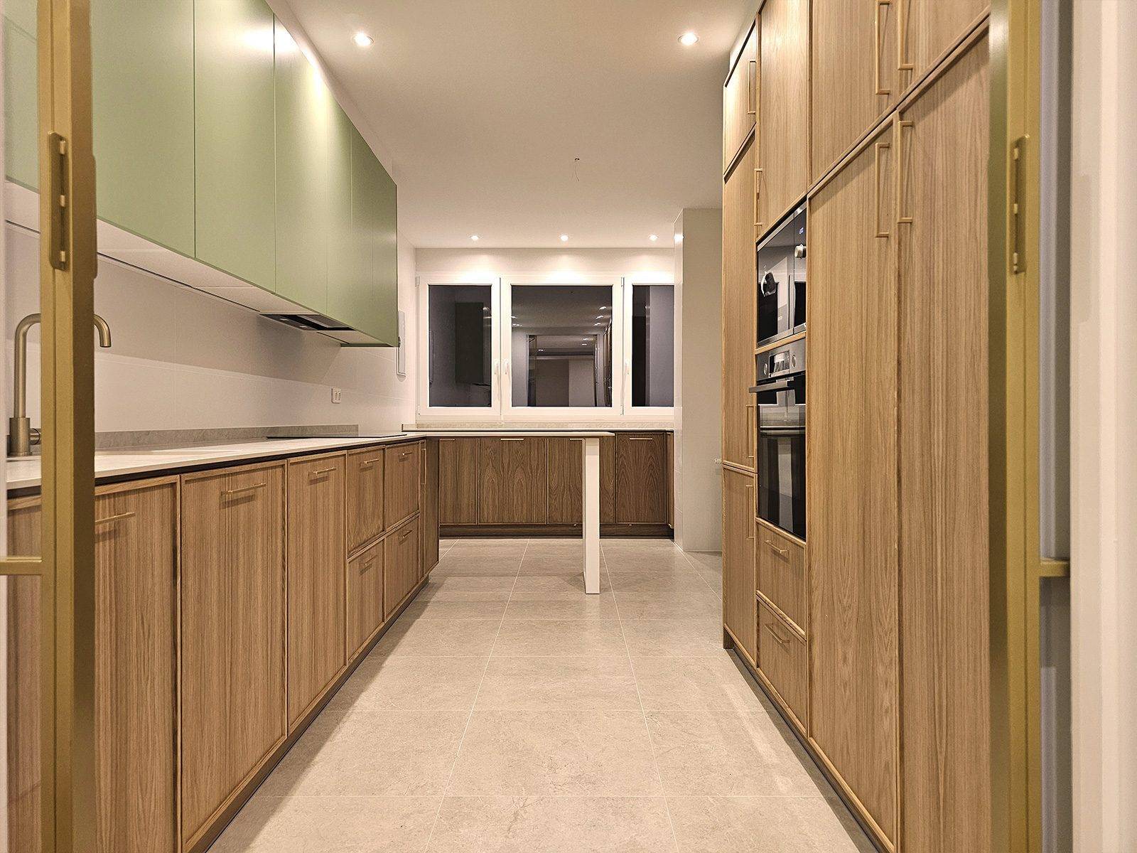 Cocina alargada con muebles bajos de madera, muebles altos verdes y columna de hornos