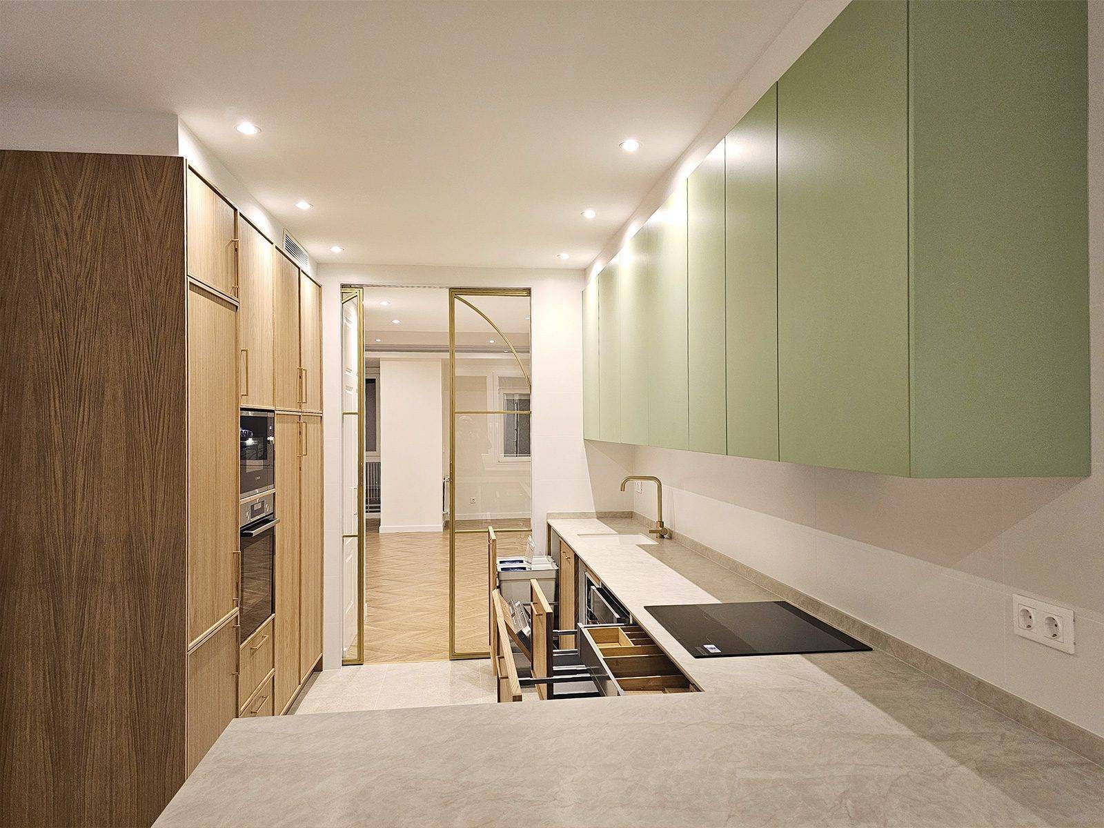 Cocina en madera y verde con placa de inducción y cajones abiertos, con la puerta de cristal al fondo