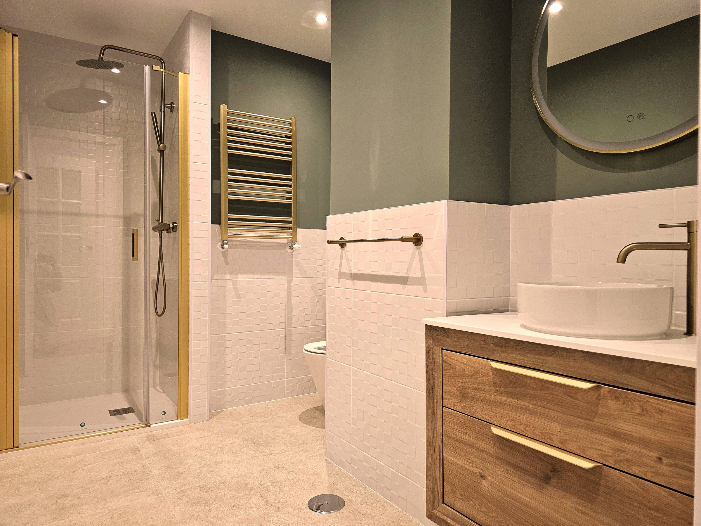 Baño con paredes en verde y azulejo blanco, lavabo sobre encimera, mueble de madera con tiradores dorados y ducha de cristal