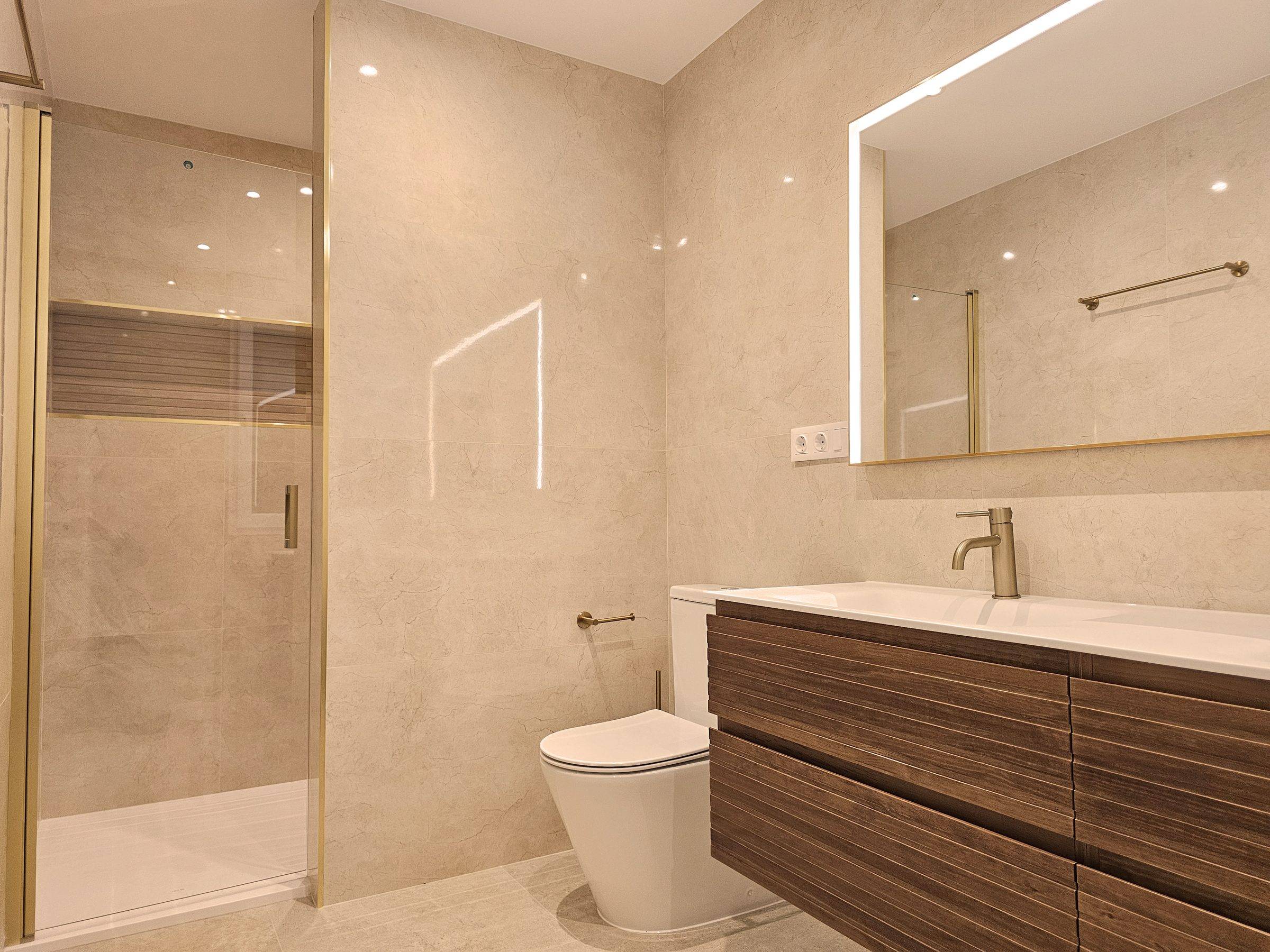Baño en tonos arena con mueble de lavabo de madera, espejo iluminado y ducha con hornacina
