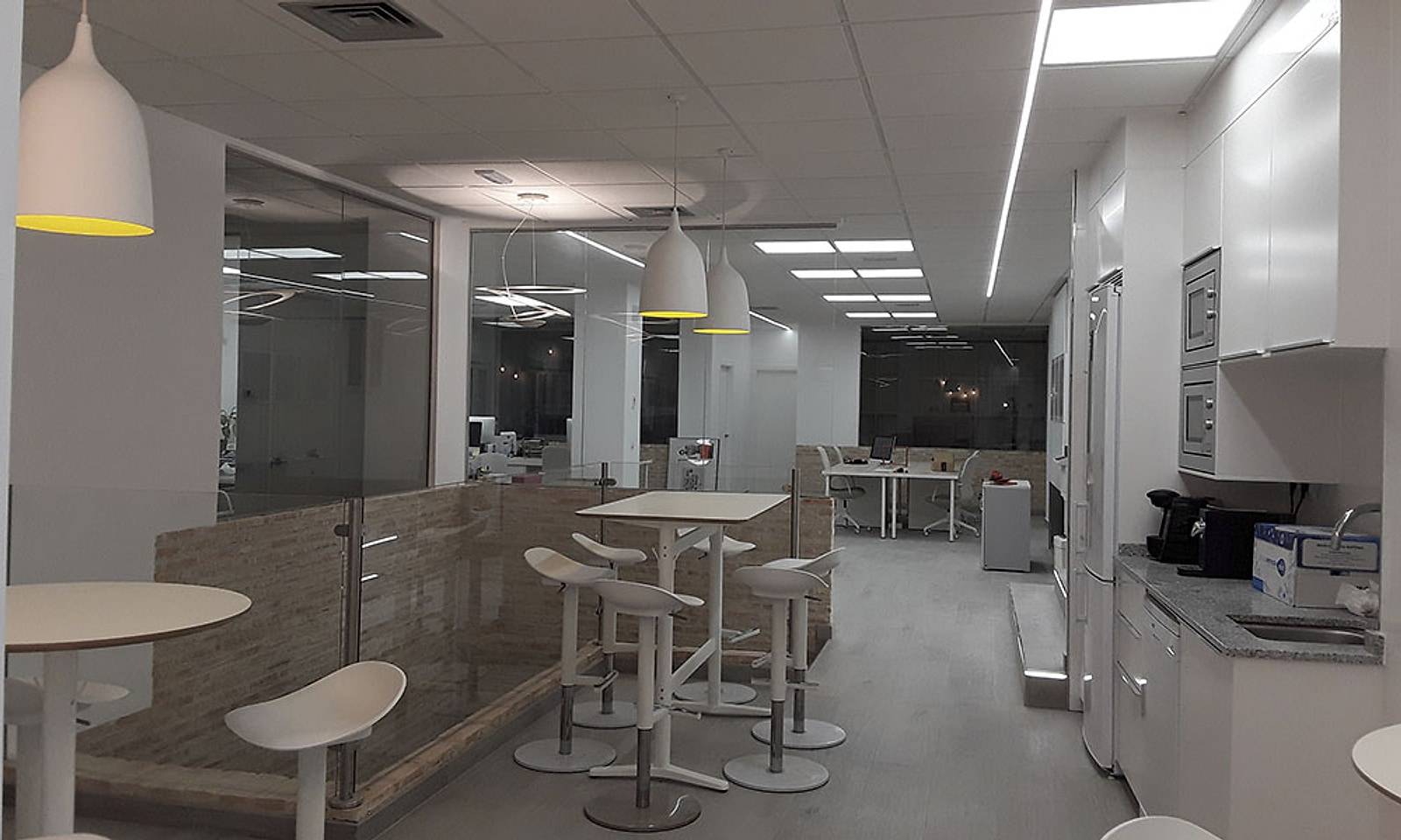 Zona de office con mesas altas, taburetes, lámparas colgantes y cocina blanca