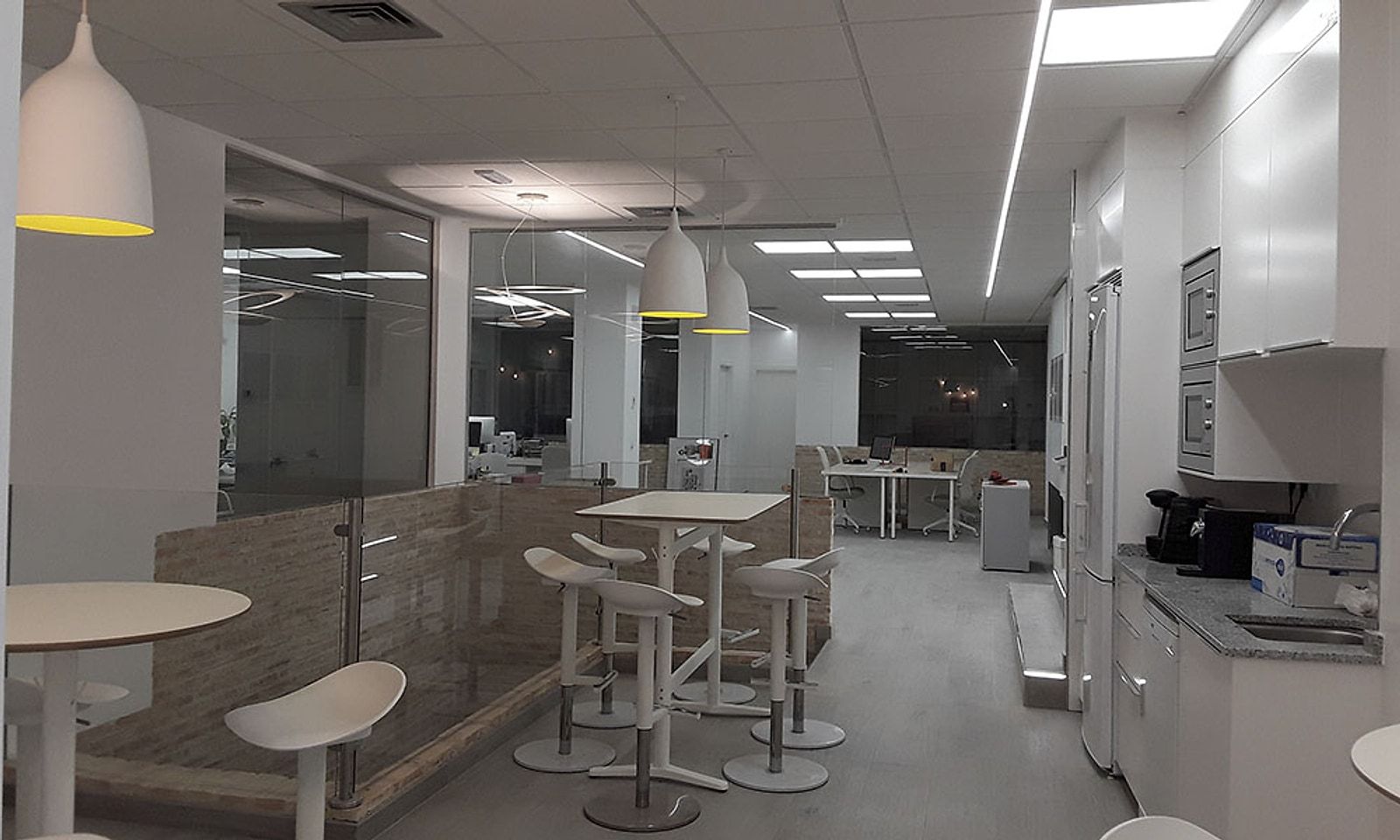 Zona de office con mesas altas, taburetes, lámparas colgantes y cocina blanca