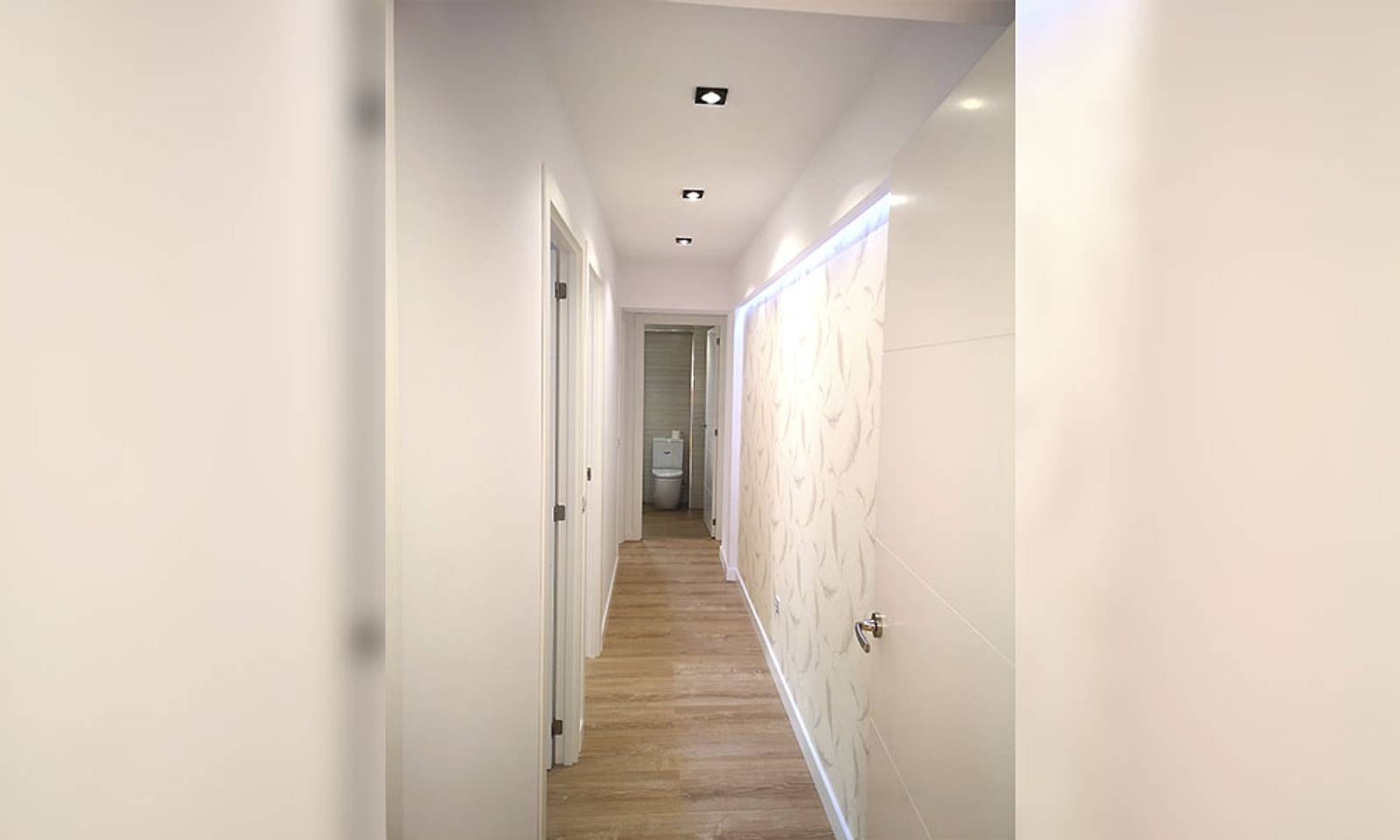 El mismo pasillo terminado, con suelo de madera, puertas blancas y una pared iluminada