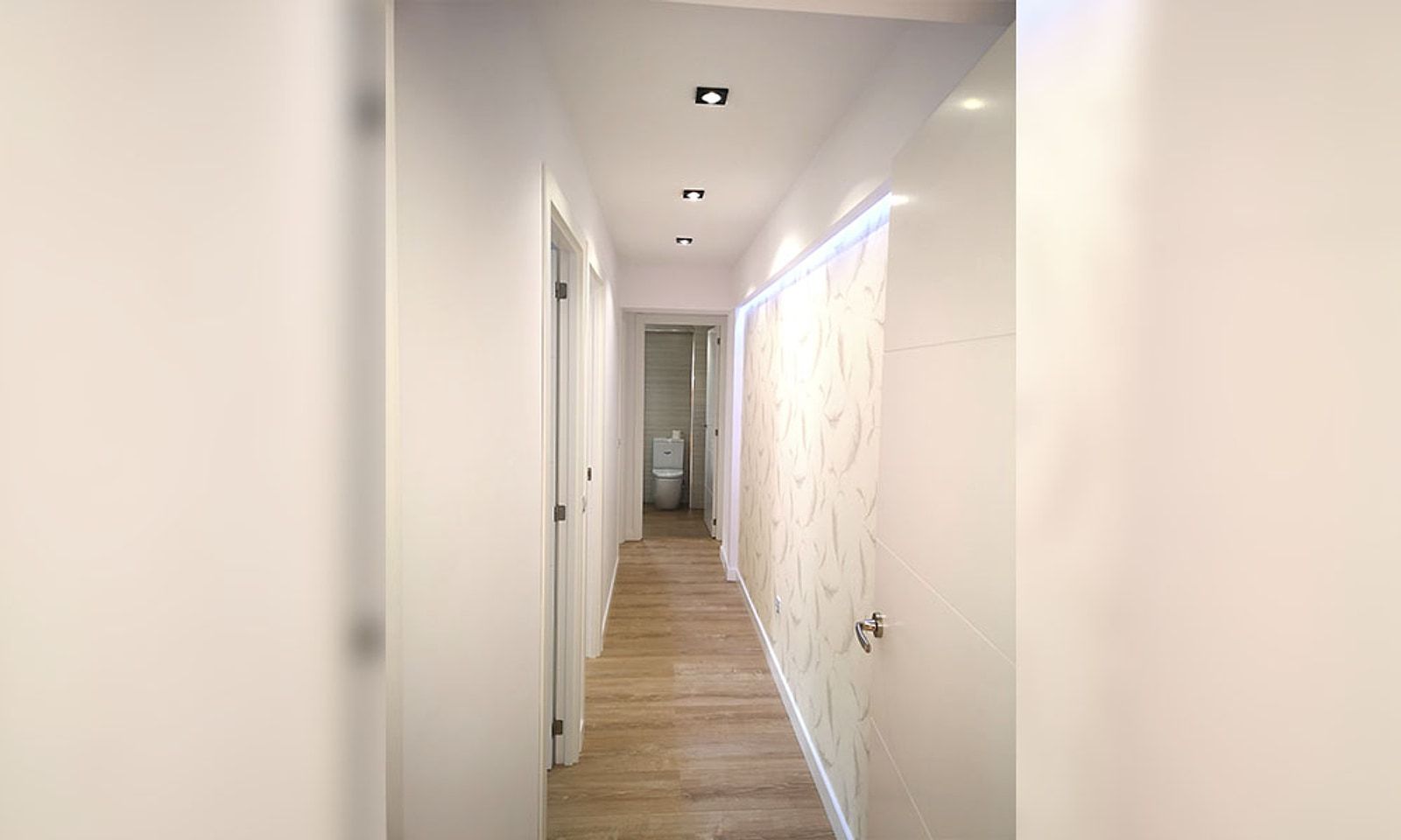 El mismo pasillo terminado, con suelo de madera, puertas blancas y una pared iluminada